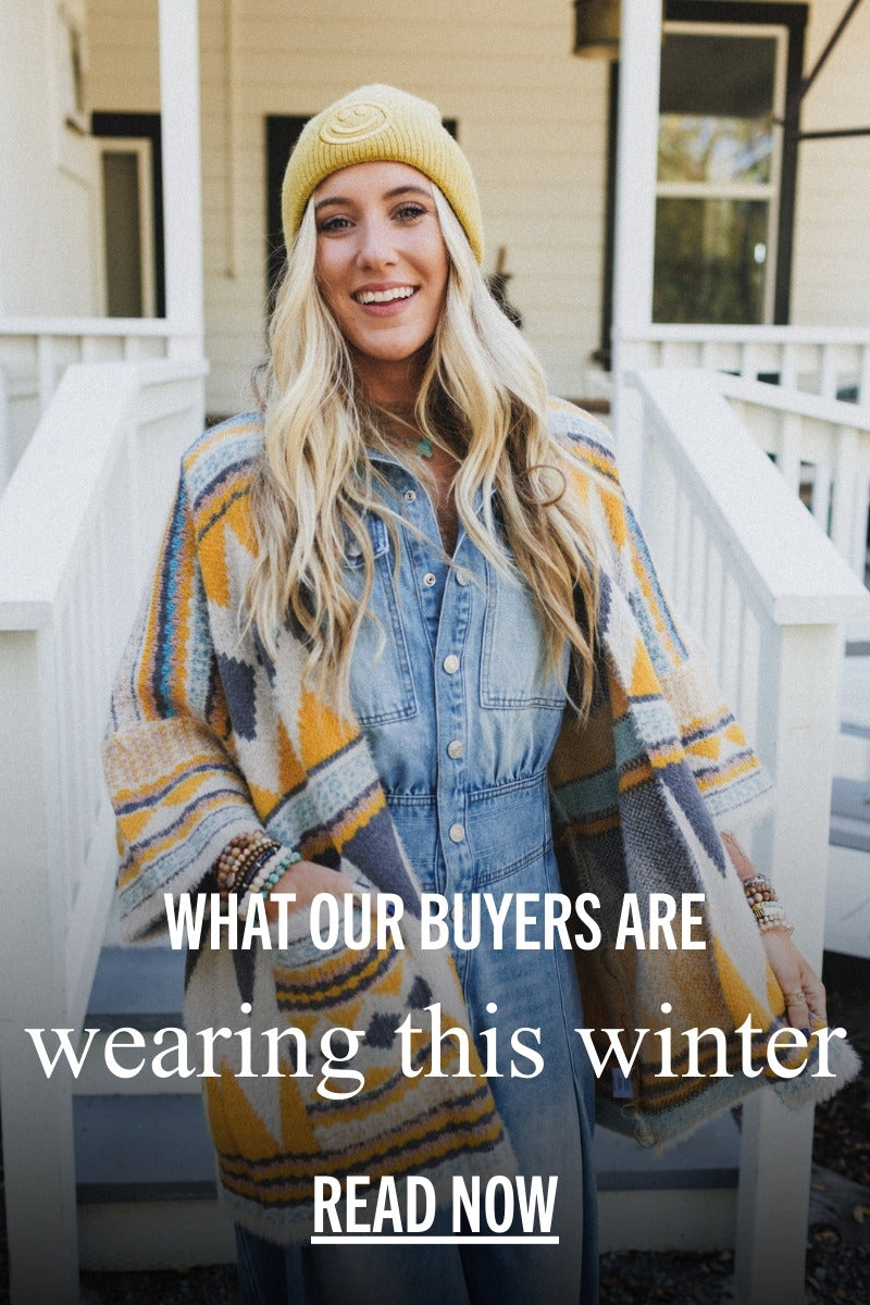 A woman wearing a yellow beanie, a denim button-down dress, and a colorful Southwestern-patterned cardigan stands smiling on a porch. This lifestyle photo introduces the Three Bird Nest blog post about what the brand’s buyers are wearing for winter.