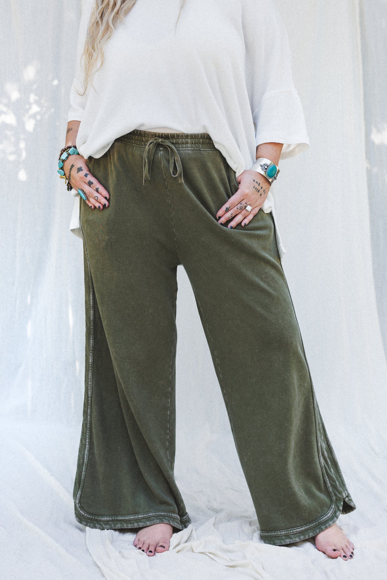 A person stands barefoot on a white fabric background, wearing the So Comfy Wide Leg Pant Cropped Length in Olive with a flowy white top. Hands with bracelets and rings are tucked into pockets, and their face is not visible.