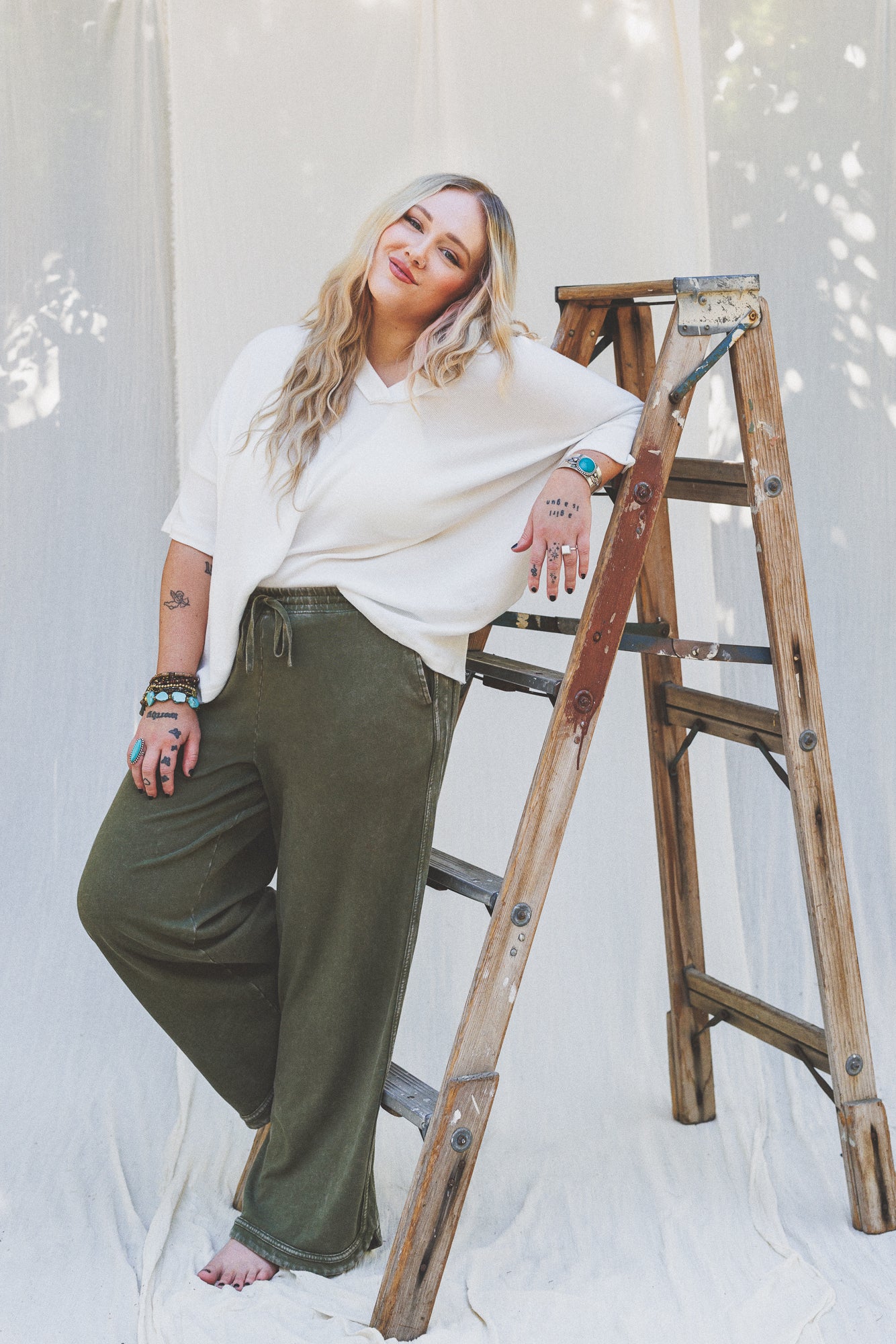 A barefoot woman with long blonde hair, wearing a white top and So Comfy Wide Leg Pant Cropped Length in olive, leans casually on a wooden ladder in front of a white fabric backdrop, smiling at the camera.