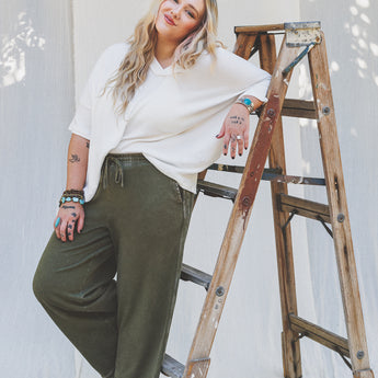 A barefoot woman with long blonde hair, wearing a white top and So Comfy Wide Leg Pant Cropped Length in olive, leans casually on a wooden ladder in front of a white fabric backdrop, smiling at the camera.