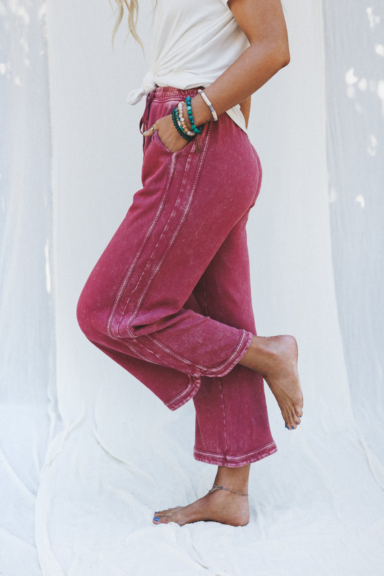 A woman stands barefoot on one leg, wearing the So Comfy Wide Leg Pant Cropped Length in Burgundy and a white shirt, accessorized with bracelets and an anklet. She poses with one hand in her pocket against a light, draped fabric background.