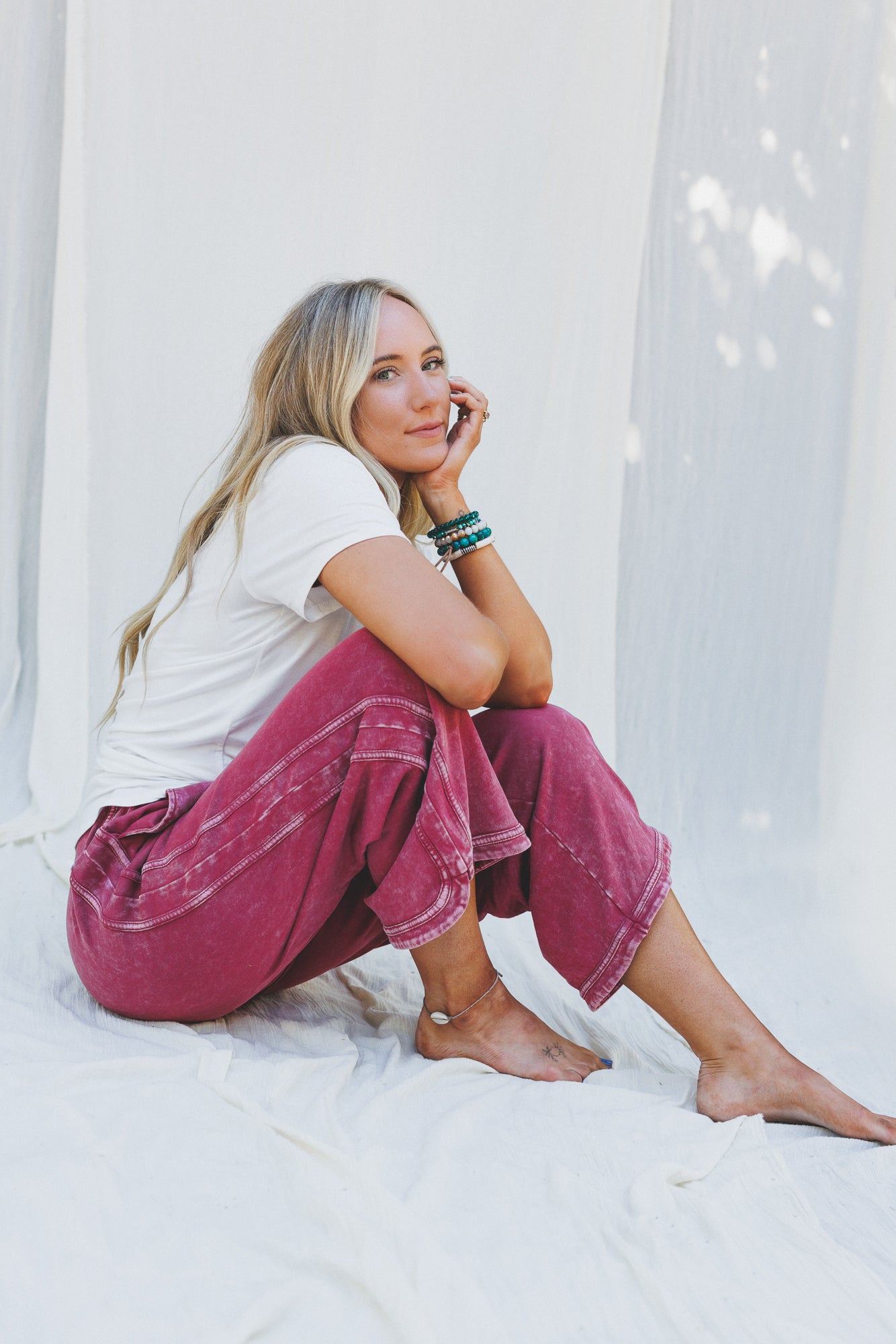 A woman with long blonde hair sits barefoot against a white fabric backdrop, wearing a white t-shirt and the So Comfy Wide Leg Pant Cropped Length in Burgundy, accessorized with bracelets, gazing relaxedly at the camera.