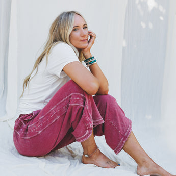 A woman with long blonde hair sits barefoot against a white fabric backdrop, wearing a white t-shirt and the So Comfy Wide Leg Pant Cropped Length in Burgundy, accessorized with bracelets, gazing relaxedly at the camera.