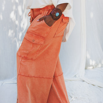A person stands barefoot on a white fabric backdrop, wearing Game On Cargo Wide Leg Pant in burnt orange with large pockets and a white top, accessorized with a wooden bracelet and beaded necklace. The setting is bright and softly lit.