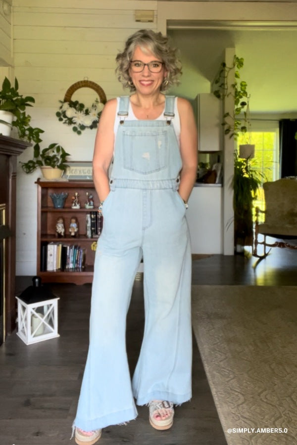 A woman with short, wavy gray hair and glasses smiles indoors with her hands in the pockets of the Milly Distressed Hem Wide Leg Overall - Denim. She pairs it with a beige top and sneakers in a boho-inspired room filled with plants and shelves.