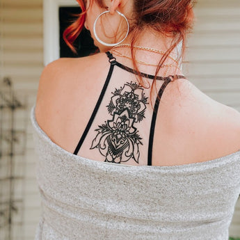 A woman with red hair in a loose bun wears hoop earrings and the Tattoo Bralette - Black under a gray cardigan off her shoulders, showing an intricate black floral tattoo on her upper back.