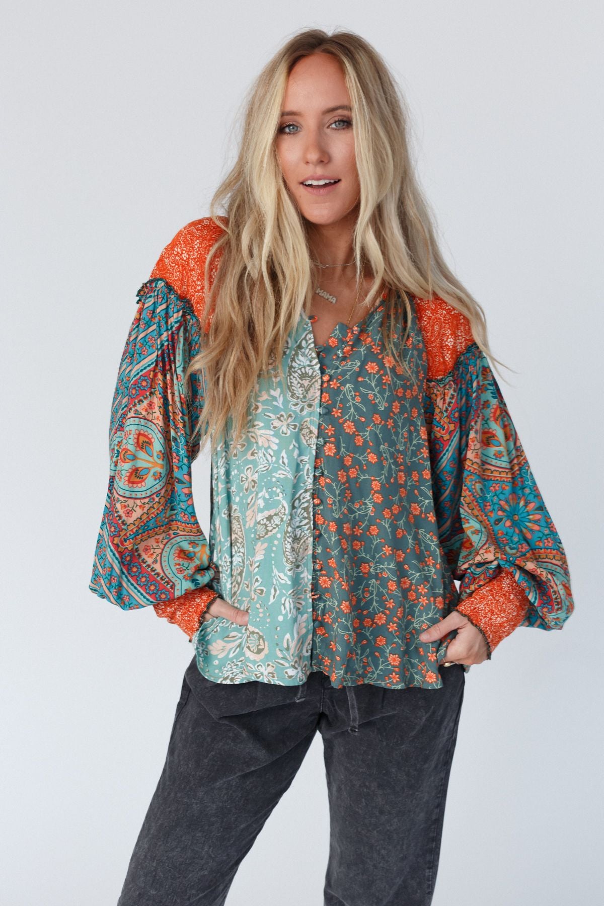 A woman with long blonde hair wears the Danielle Mixed Floral Printed Top - Mint, featuring mixed patterns in teal, orange, and red, paired with black washed jeans against a plain light background.