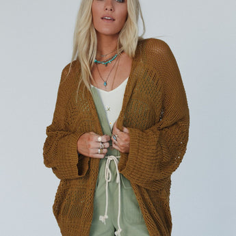 A woman with long blonde hair wears the Brenden Knit Netted Cardigan in brown over a white top and green shorts, accessorized with layered necklaces and rings, standing against a plain light background.