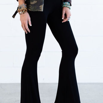 Wearing Cher Solid Raw Edge Flare Pant in black, a person stands against a white wall. The photo is cropped to display only the flared pants and lower body, styled with a tied white shirt, camouflage jacket, and bracelets.
