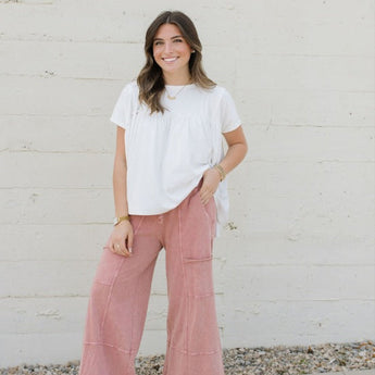 A smiling woman stands before a light wall, wearing the Lucky One Wide Leg Pant in Mauve with a white short-sleeve top, white sandals, and accessories for a boho style. She has long brown hair styled naturally.