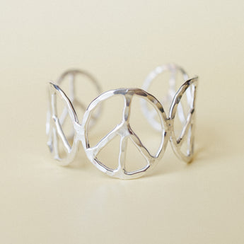 A wide silver cuff bracelet with repeating peace sign cutouts arranged in an open, sculptural band — shot close up against a warm cream background with soft, even light.