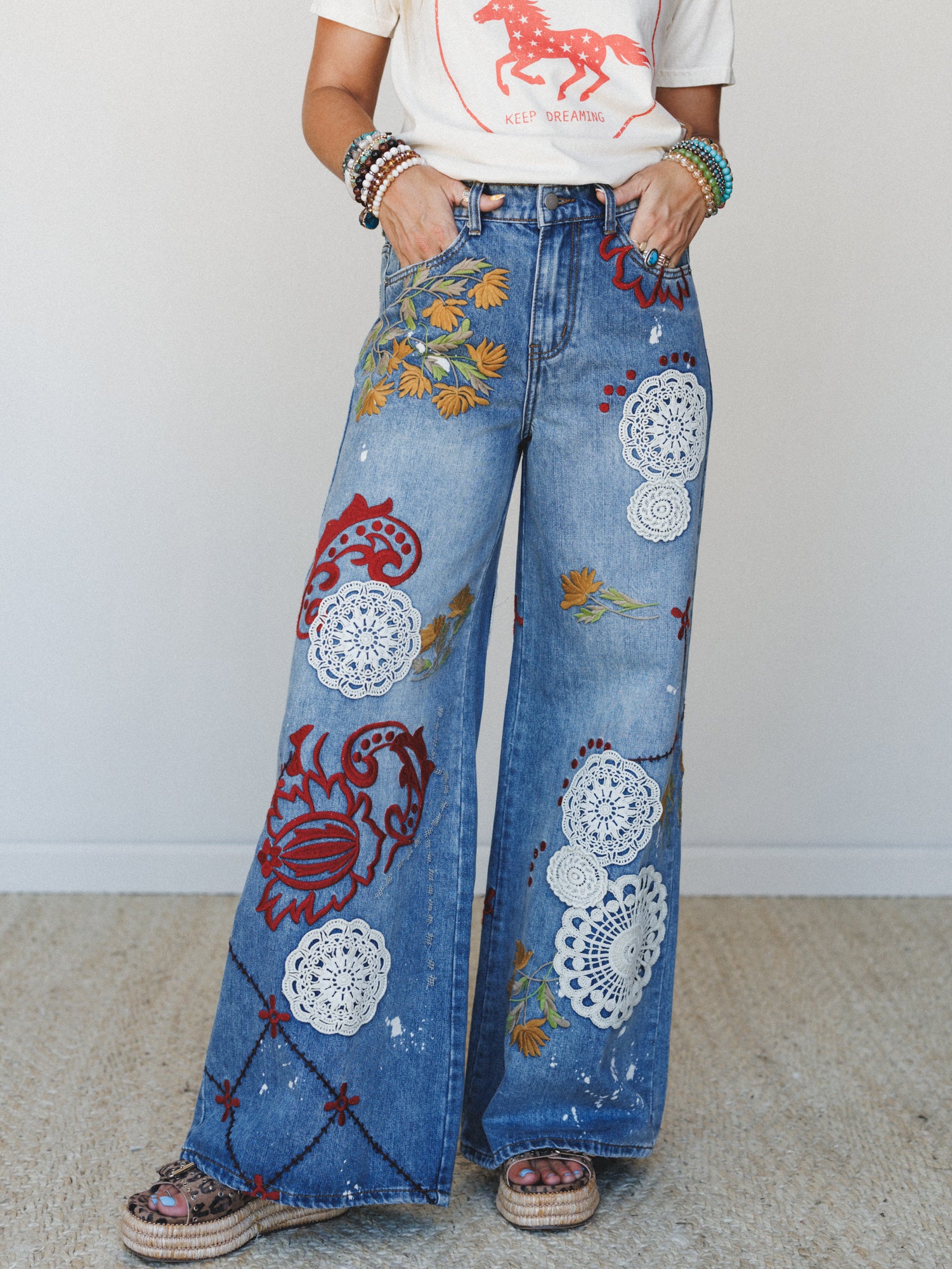 Wide-leg embellished jeans covered in crochet doily appliqués, floral embroidery, and red felt-like patchwork motifs, styled with a graphic tee and stacked bracelets — the kind of jeans that tell a whole story before you say a word.