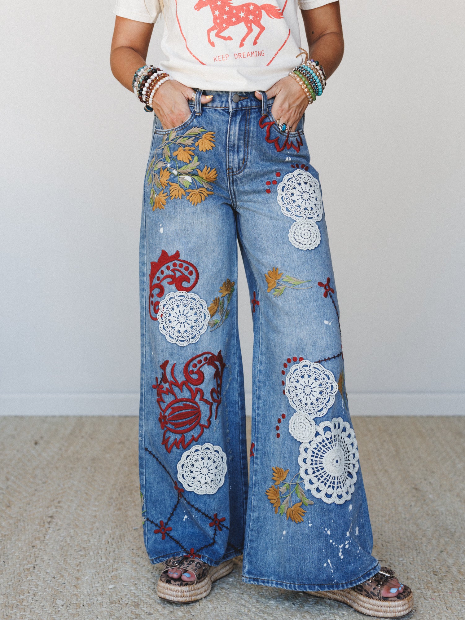 Wide-leg denim jeans covered in crochet doily appliqués, floral embroidery, and bold red folk-art motifs — styled with a graphic tee and stacked bracelets for a lived-in, expressive boho look.
