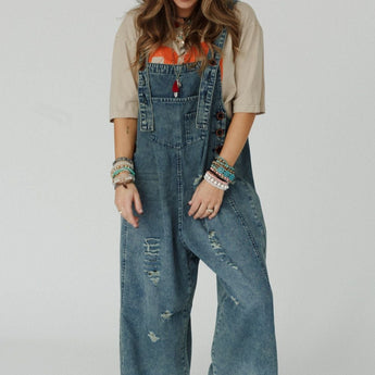 A woman smiles, looking down. She wears a beige T-shirt and the High Hopes Slouchy Denim Overalls - Blue with adjustable straps, paired with white sneakers and colorful bracelets. Her wavy hair is loose against a light gray backdrop.