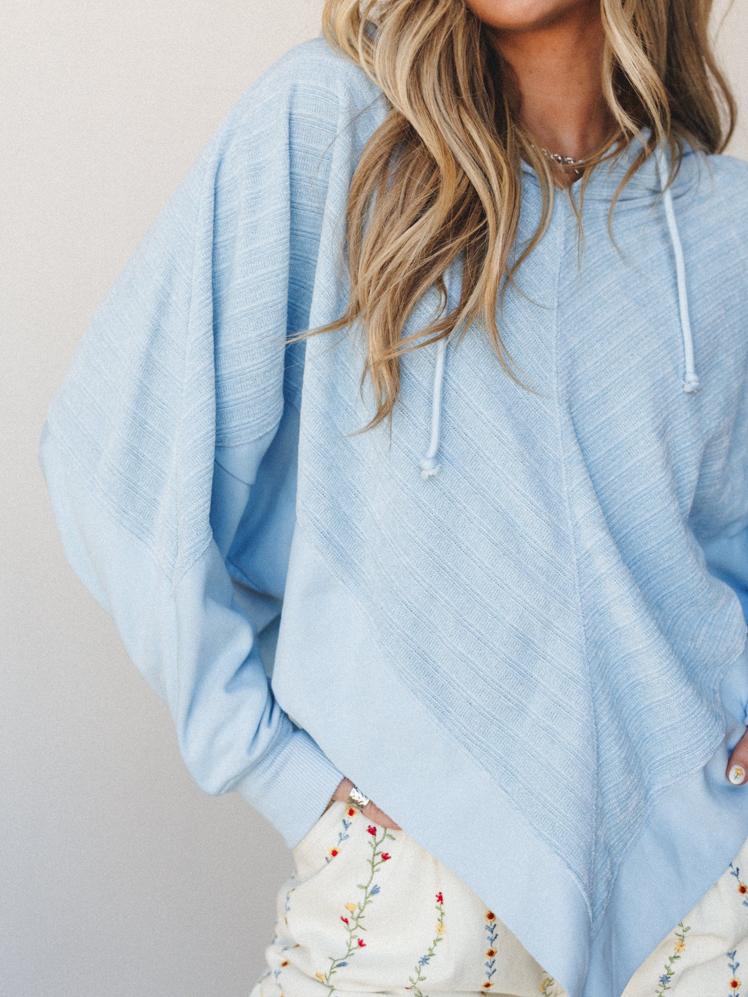 A soft periwinkle hoodie with oversized batwing sleeves and diagonal ribbed texture, styled over a cream floral embroidered bottom — the kind of outfit that feels like a good-mood day without trying too hard.
