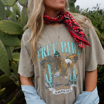 A woman stands outdoors in front of cacti, wearing the Fly Free Graphic Tee - Mocha, a red bandana around her neck, layered necklaces, and a light denim jacket draped off her shoulders.
