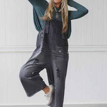 A smiling woman playfully poses on one foot in a black cap, teal long-sleeve shirt, and High Hopes Slouchy Denim Overalls - Black, featuring distressed details. She completes the look with white sneakers against a white paneled wall.