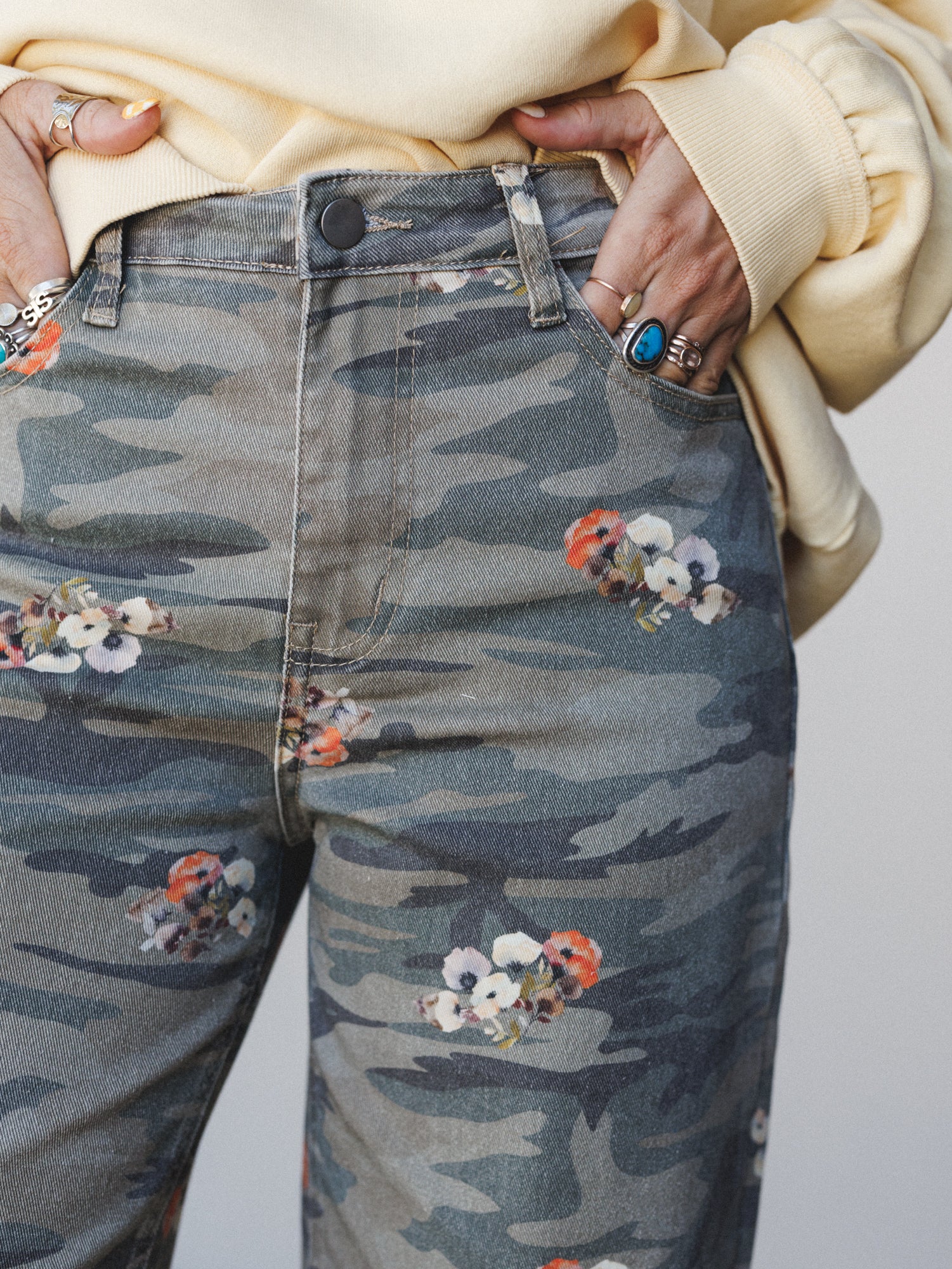 A close-up of camo-print denim jeans scattered with small floral clusters, styled with a cream sweatshirt and layered silver and turquoise rings — the kind of outfit that feels effortlessly collected without trying too hard.