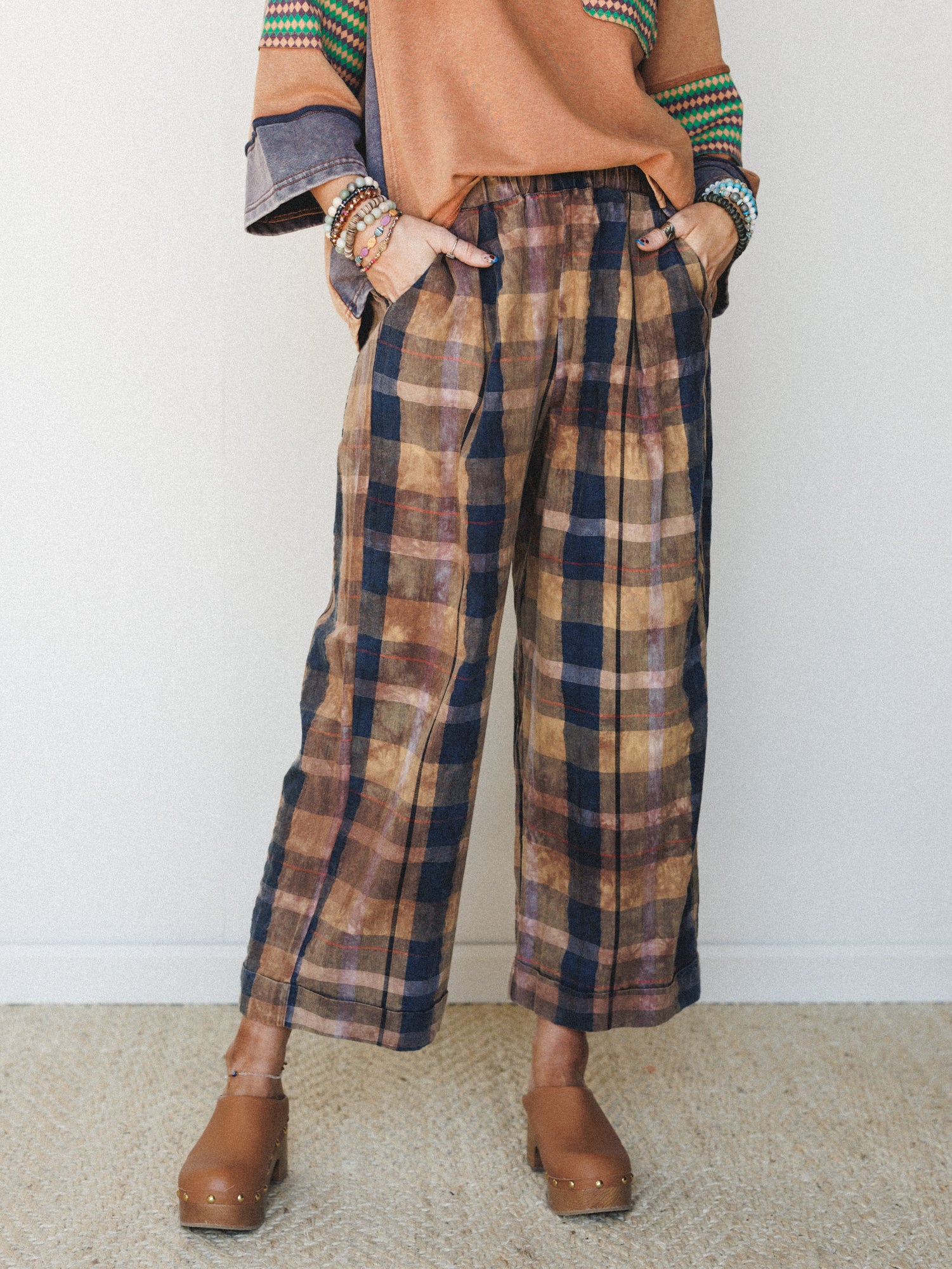 Wide-leg plaid pants in a washed brown and navy palette styled with a rust sweatshirt and wooden clogs — the kind of effortlessly collected outfit that looks like it came together on purpose.