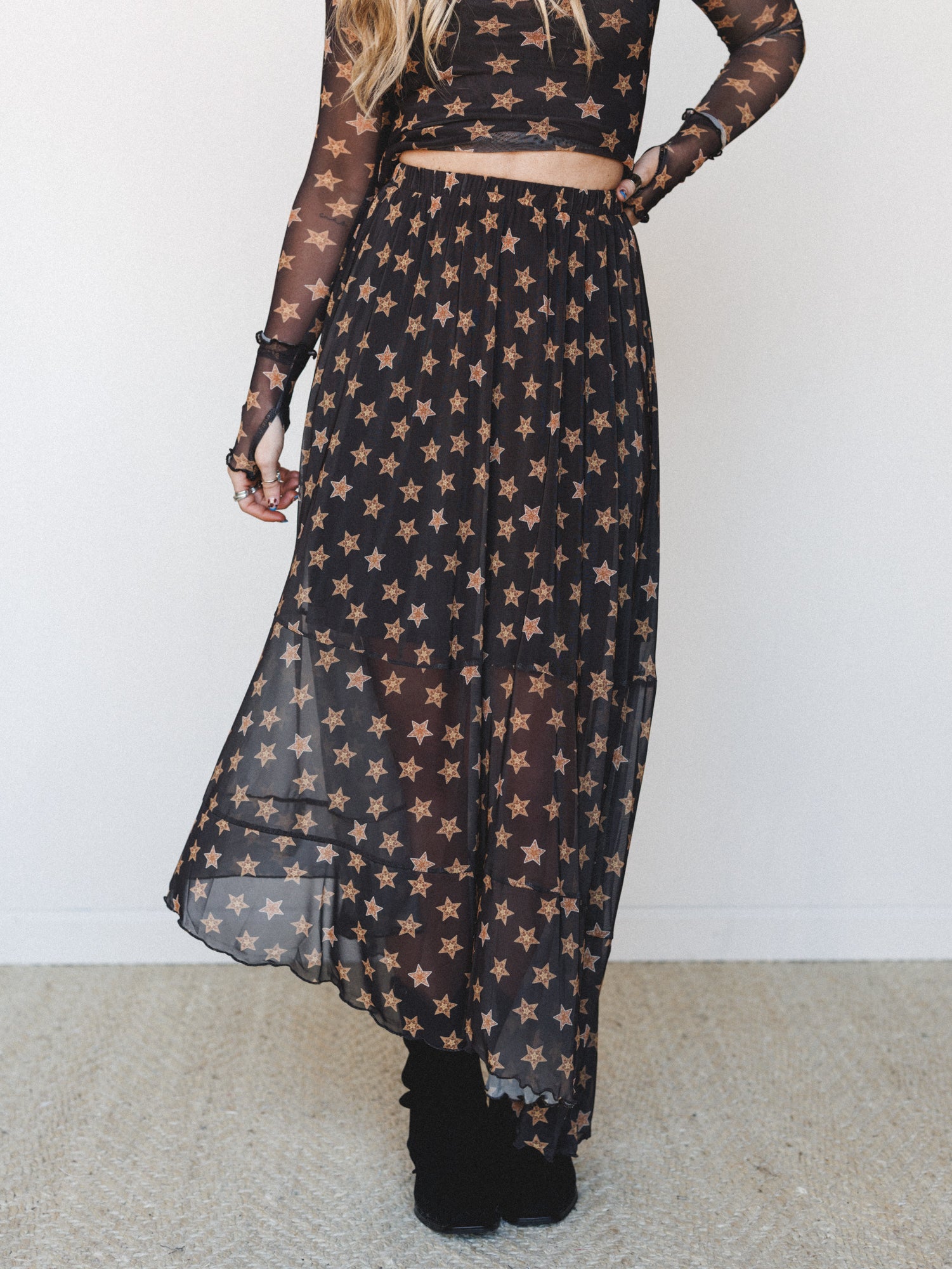 A sheer black maxi skirt covered in warm golden star print floats around the model's ankles, paired with a matching long-sleeve mesh crop top and black ankle boots — celestial and a little dramatic in the best way.