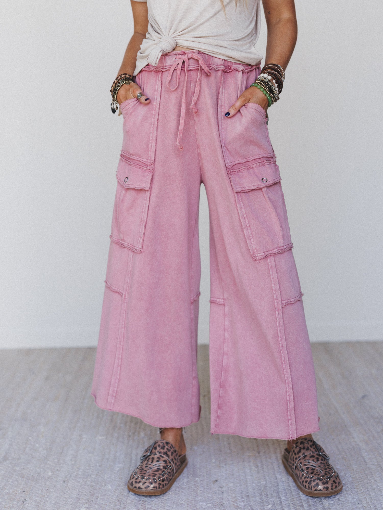 Person wearing Game On Cargo Wide Leg Pant in burgundy with a drawstring stretch waist, paired with a knotted white t-shirt, animal-print slip-ons, and layered bracelets for a comfy boho look against a plain background.
