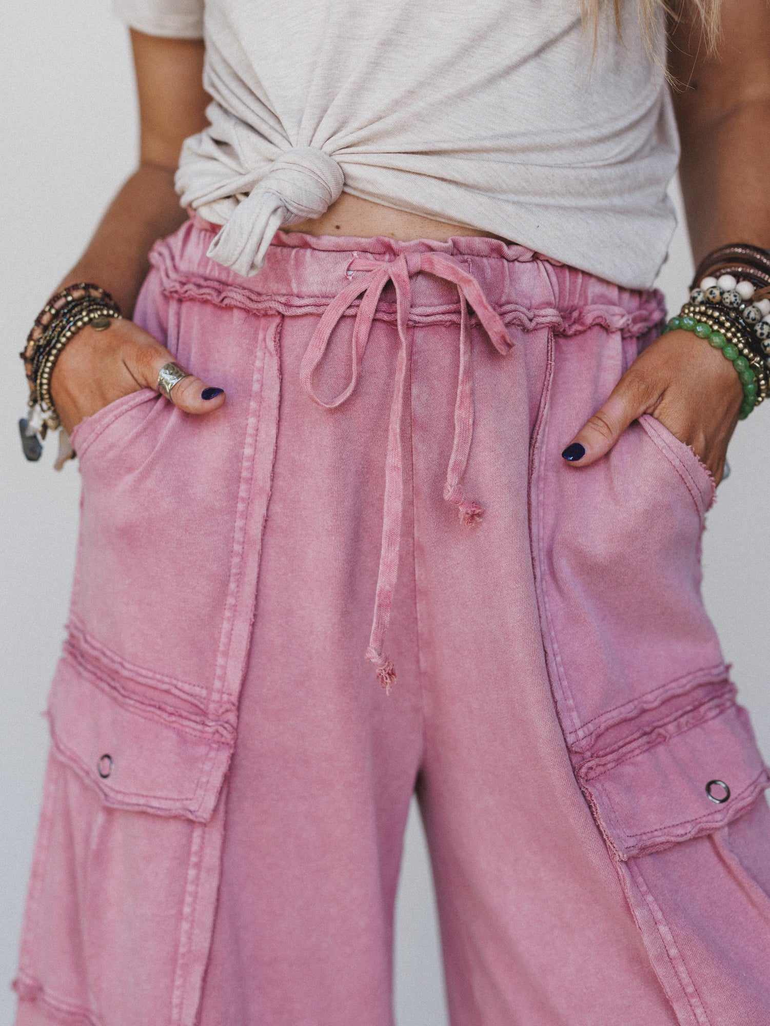 A person wears the Game On Cargo Wide Leg Pant in burgundy, styled with a knotted beige tee, hands in pockets. They complete their comfy look with several beaded bracelets and rings on both hands.