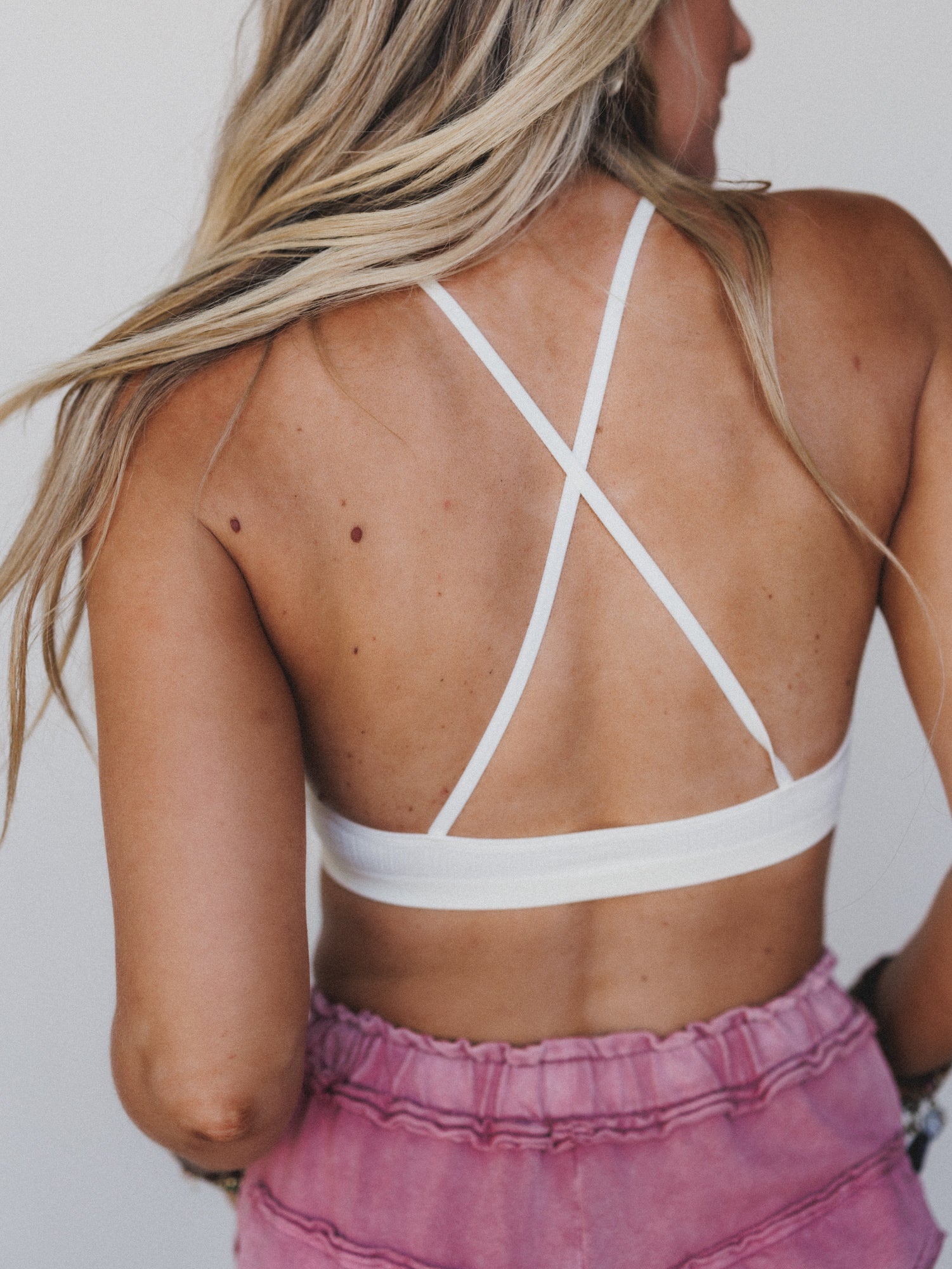 A woman with long blonde hair is shown from the back, wearing the Crochet High Neck Bralette in ivory and high-waisted pink shorts. Her loose hair cascades down, revealing sun-kissed skin with freckles and moles.