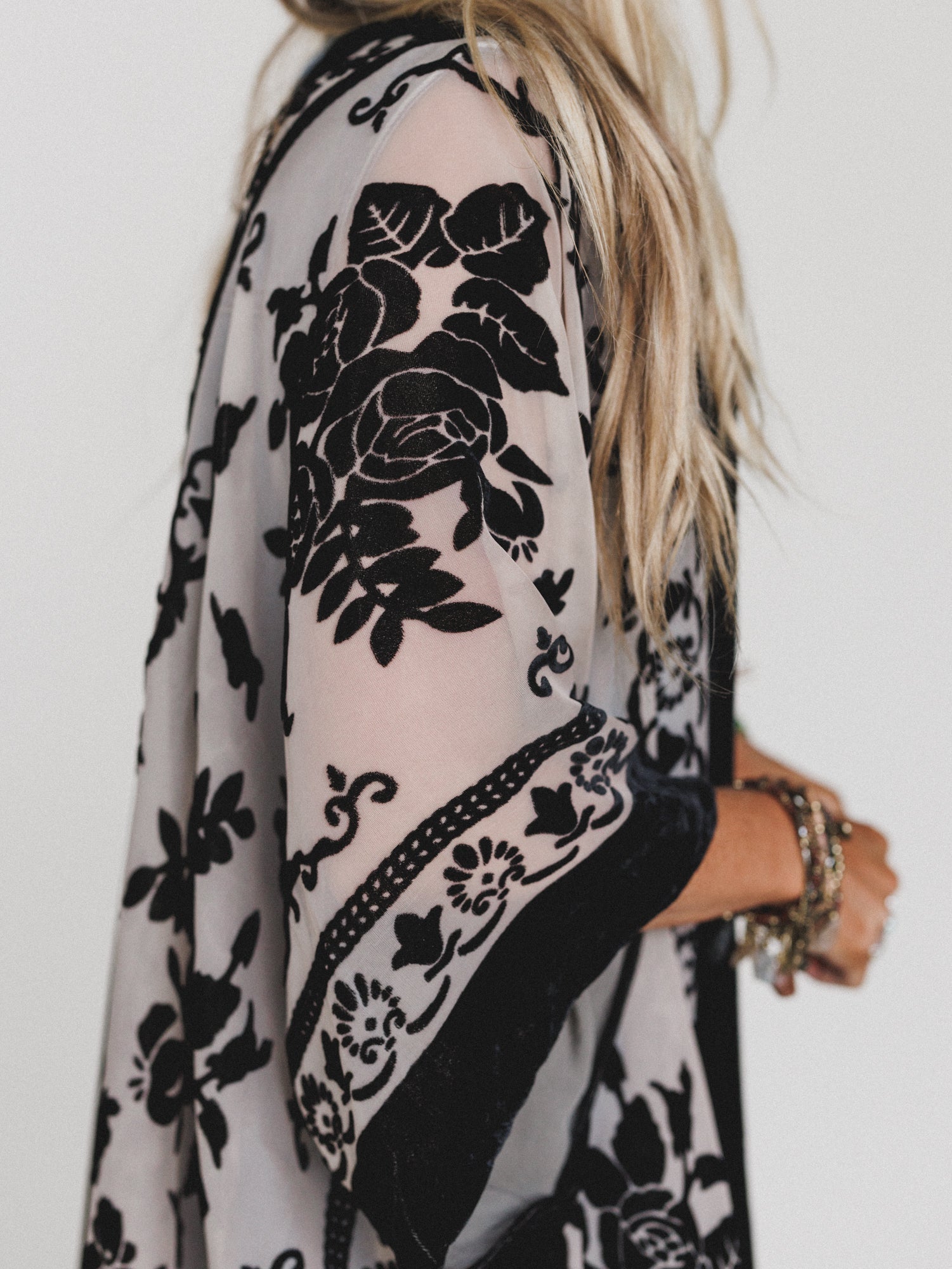 A woman with long blonde hair wears the Crushin' On You Velvet Kimono in black. Shown from the side, she displays layered bracelets on her wrist against a plain white background.