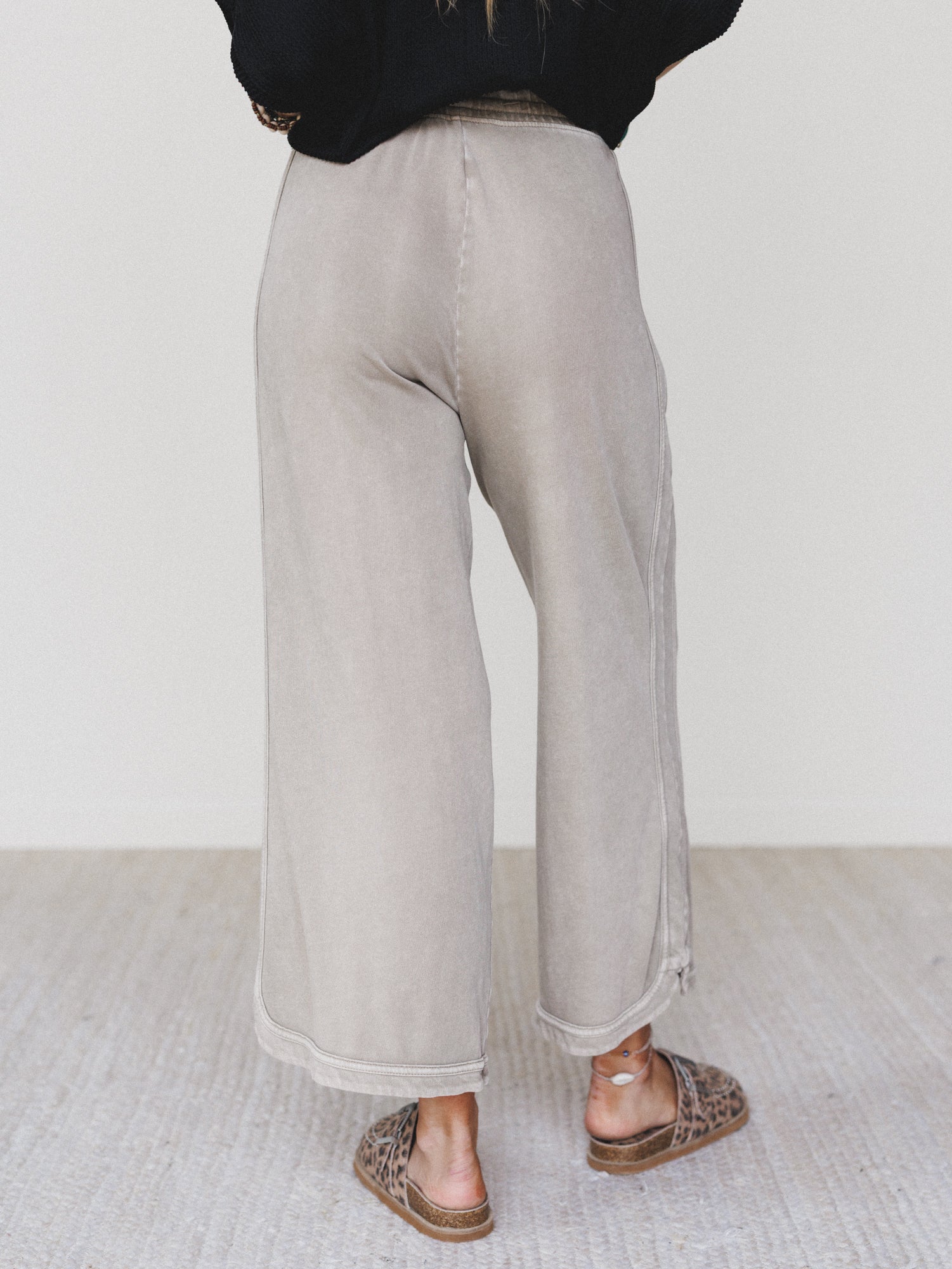 Wearing the So Comfy Wide Leg Pant Cropped Length in Mocha, featuring a mineral wash, high-rise elastic waist, and wide leg silhouette, paired with brown woven slip-on shoes on a light textured floor. Upper body not visible; hands on hips.