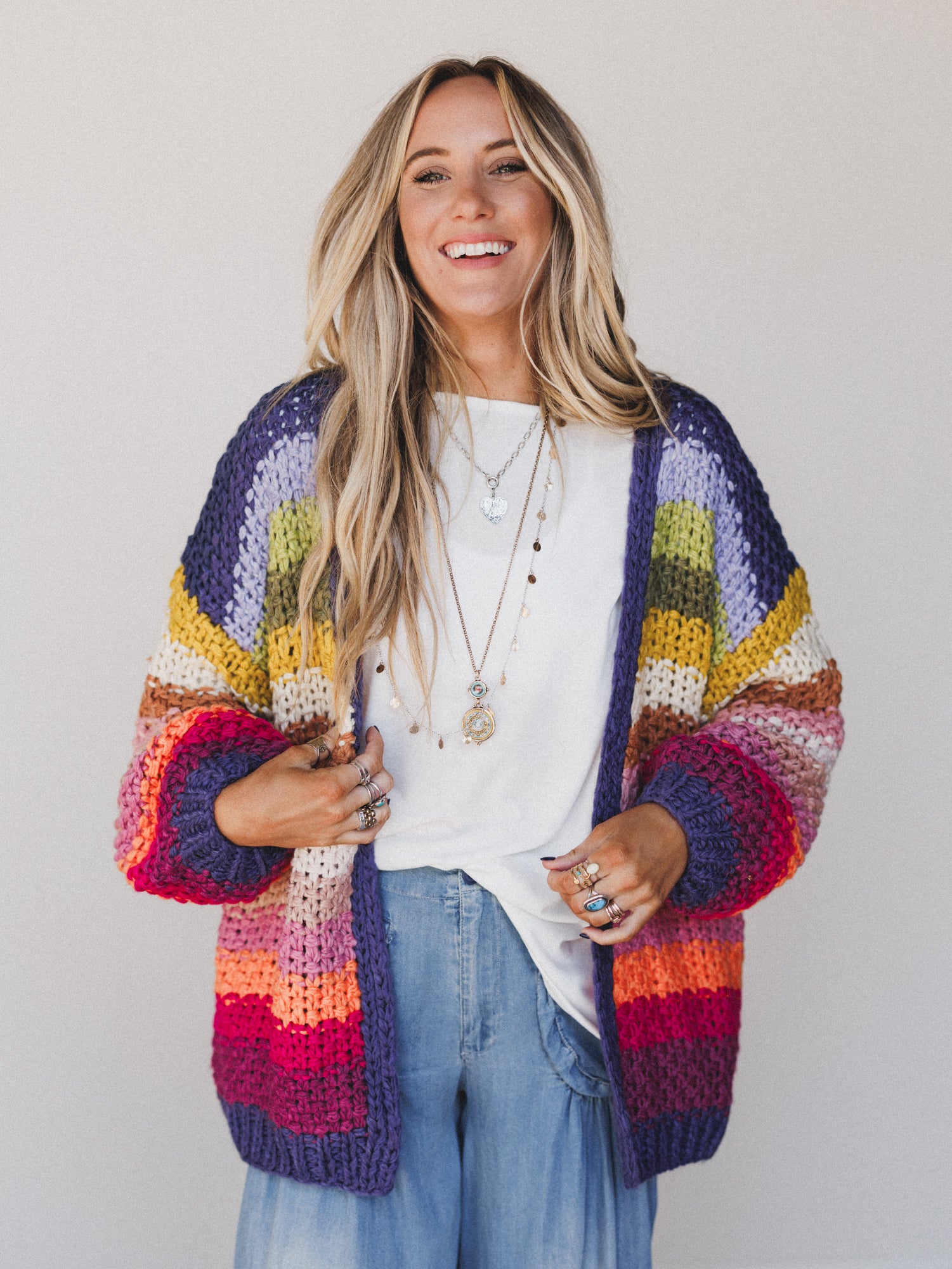 A smiling woman with long blonde hair wears the Wrapped up in Color Knit Cardigan - Rainbow, an open-front boho sweater, over a white T-shirt and loose blue jeans, styled with layered necklaces and rings against a plain light background.
