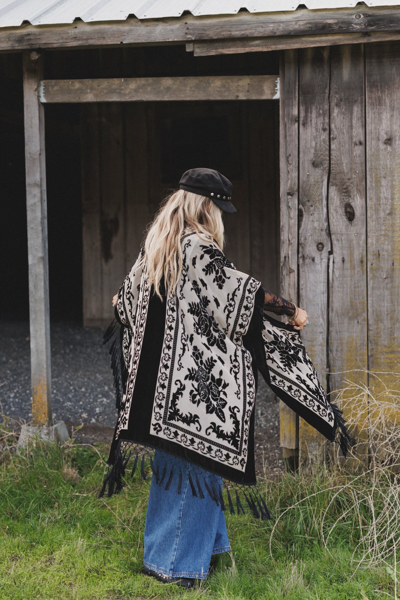 A person with long blonde hair, a black hat, the *Crushin' On You Velvet Kimono - Black*, and wide-leg jeans stands near an old wooden building with weathered walls and a metal roof.