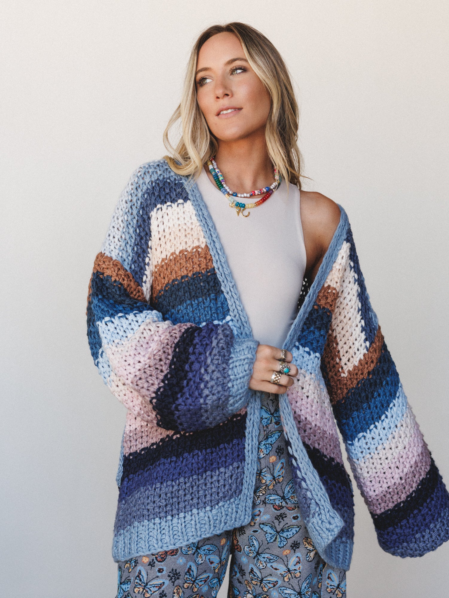 A relaxed, dreamy shot of a model wearing an oversized chunky knit stripe cardigan in a soft palette of blues, lavender, caramel, and cream — layered over a white tank and butterfly print pants, with stacked beaded necklaces that feel like a summer m