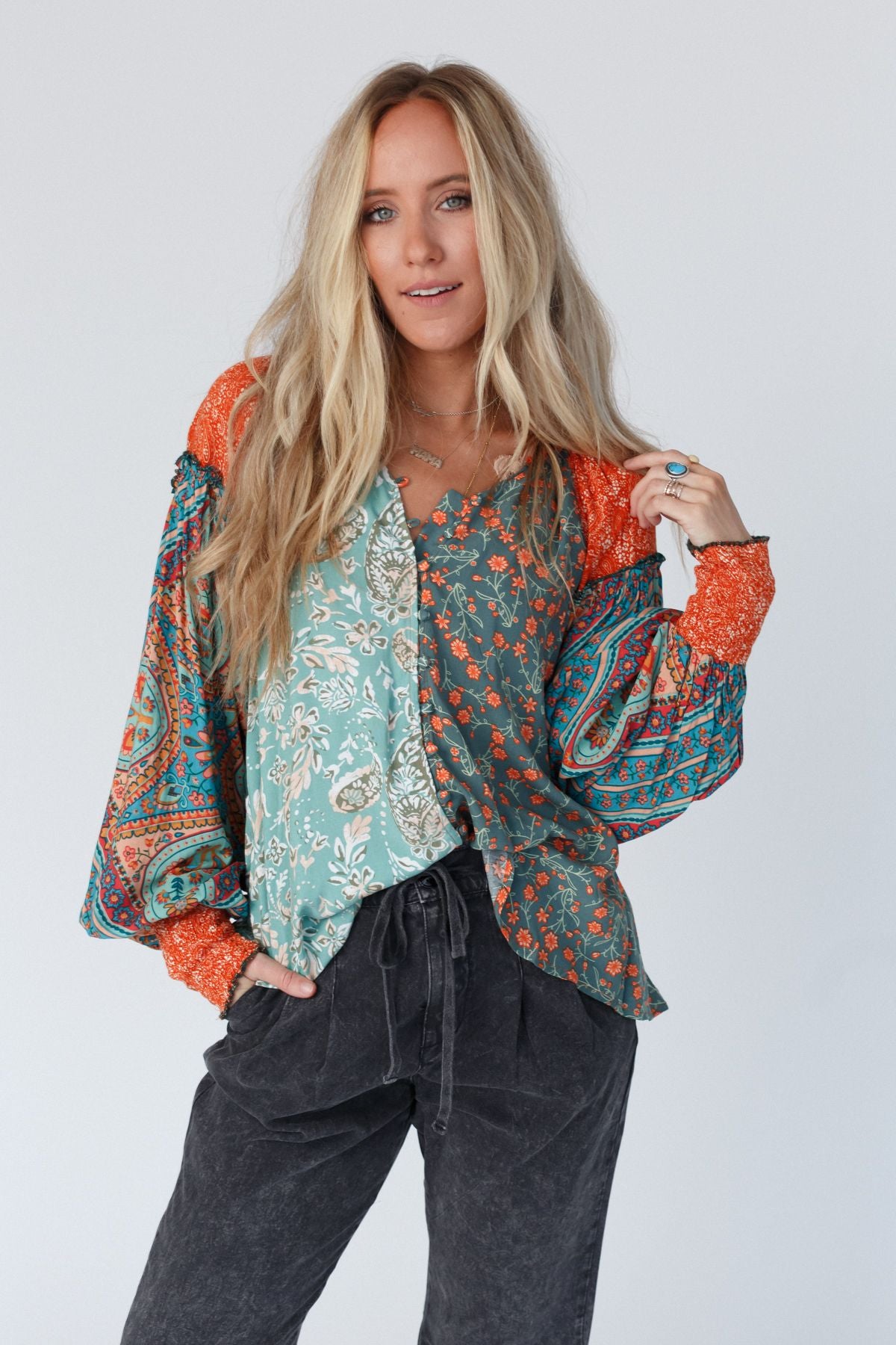 A woman with long blonde hair wears the Danielle Mixed Floral Printed Top - Mint, paired with loose dark pants. She stands against a plain light background, smiling slightly with one hand near her hair.