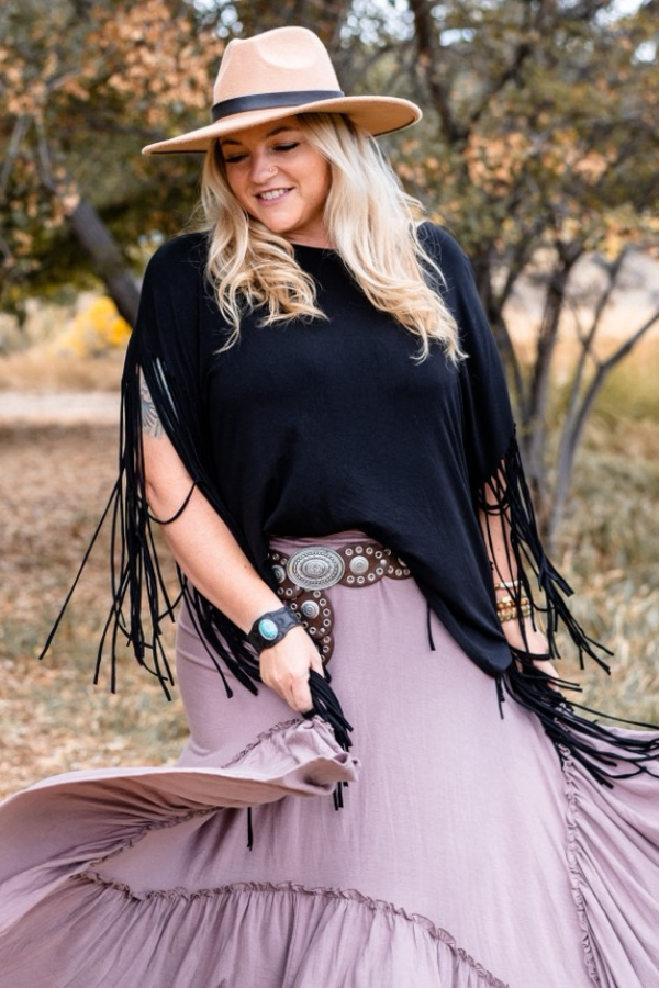 A smiling woman wears The Nest Showstopper Fringe Top in black, paired with a tan hat and mauve skirt. She twirls outdoors on an autumn path, holding her skirt and showing off a decorative belt among the trees.