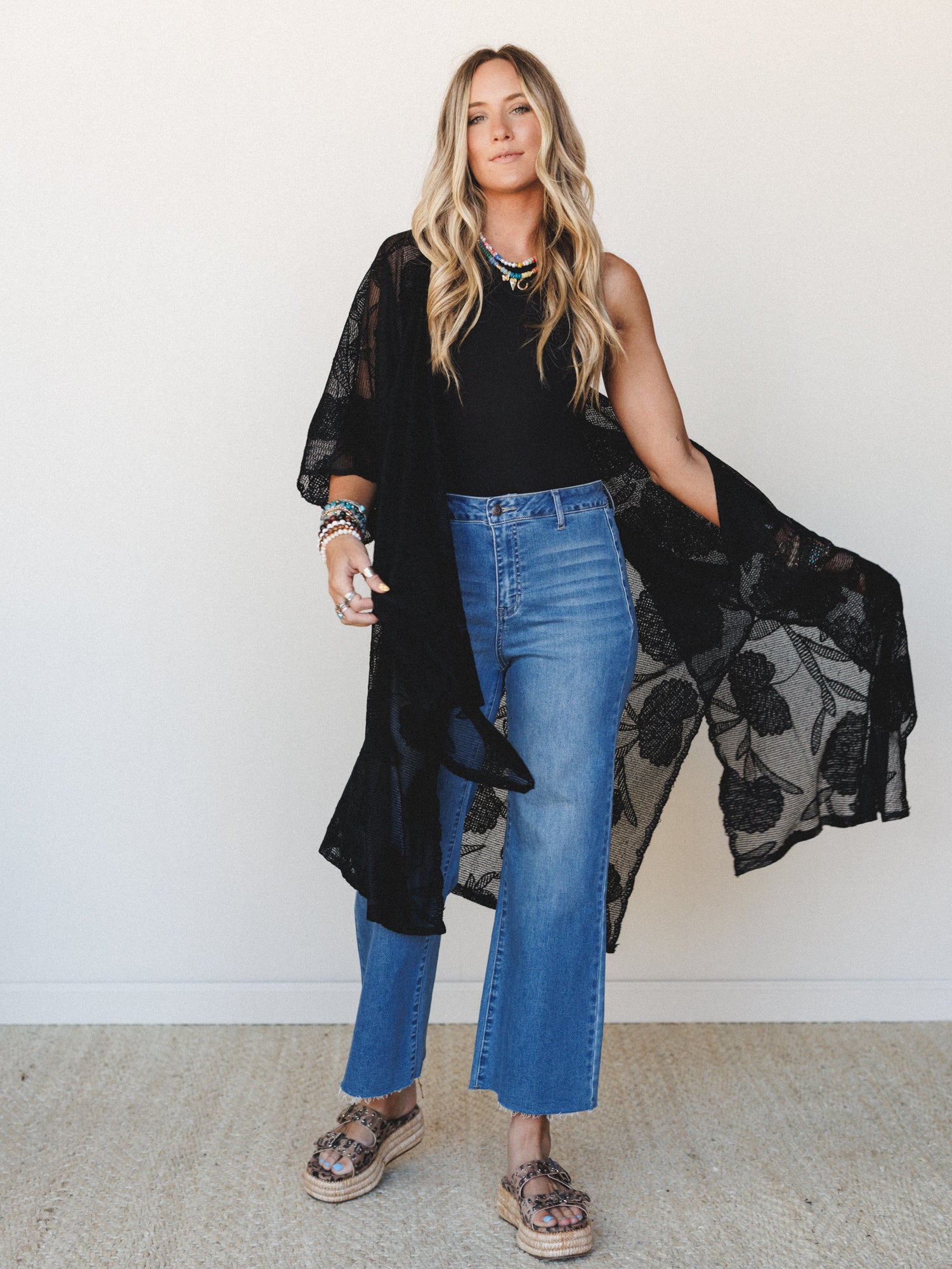 A relaxed full-body shot of a model layering a sheer black floral lace kimono over a black sleeveless top and cropped wide-leg jeans, styled with platform espadrille sandals and stacked jewelry — effortlessly boho with a cool-girl edge.