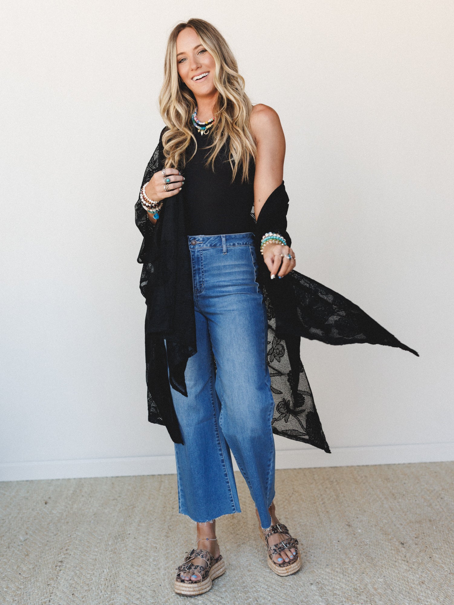 A smiling blonde model layered in a black lace-print duster over a black sleeveless top and cropped wide-leg jeans, styled with colorful beaded jewelry and espadrille platform sandals — the kind of outfit that looks effortless but definitely took thr