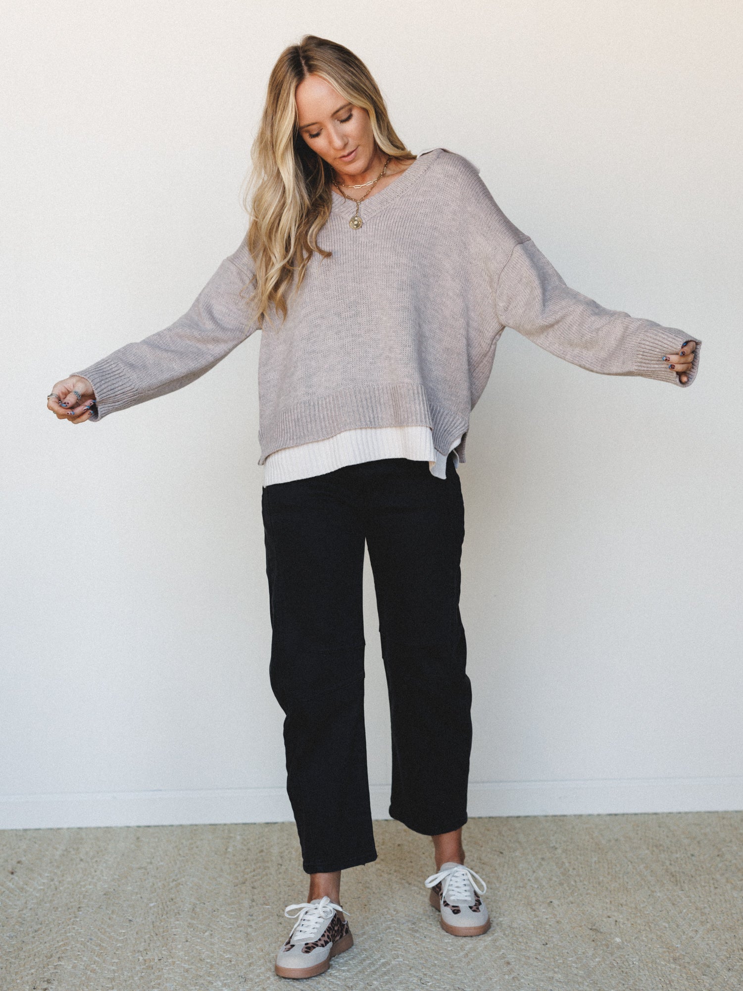 An oversized v-neck knit sweater layered over a white long-sleeve top, paired with straight-leg black pants and leopard-print sneakers — the kind of effortless, put-together look that feels like it took five minutes but somehow looks intentional.