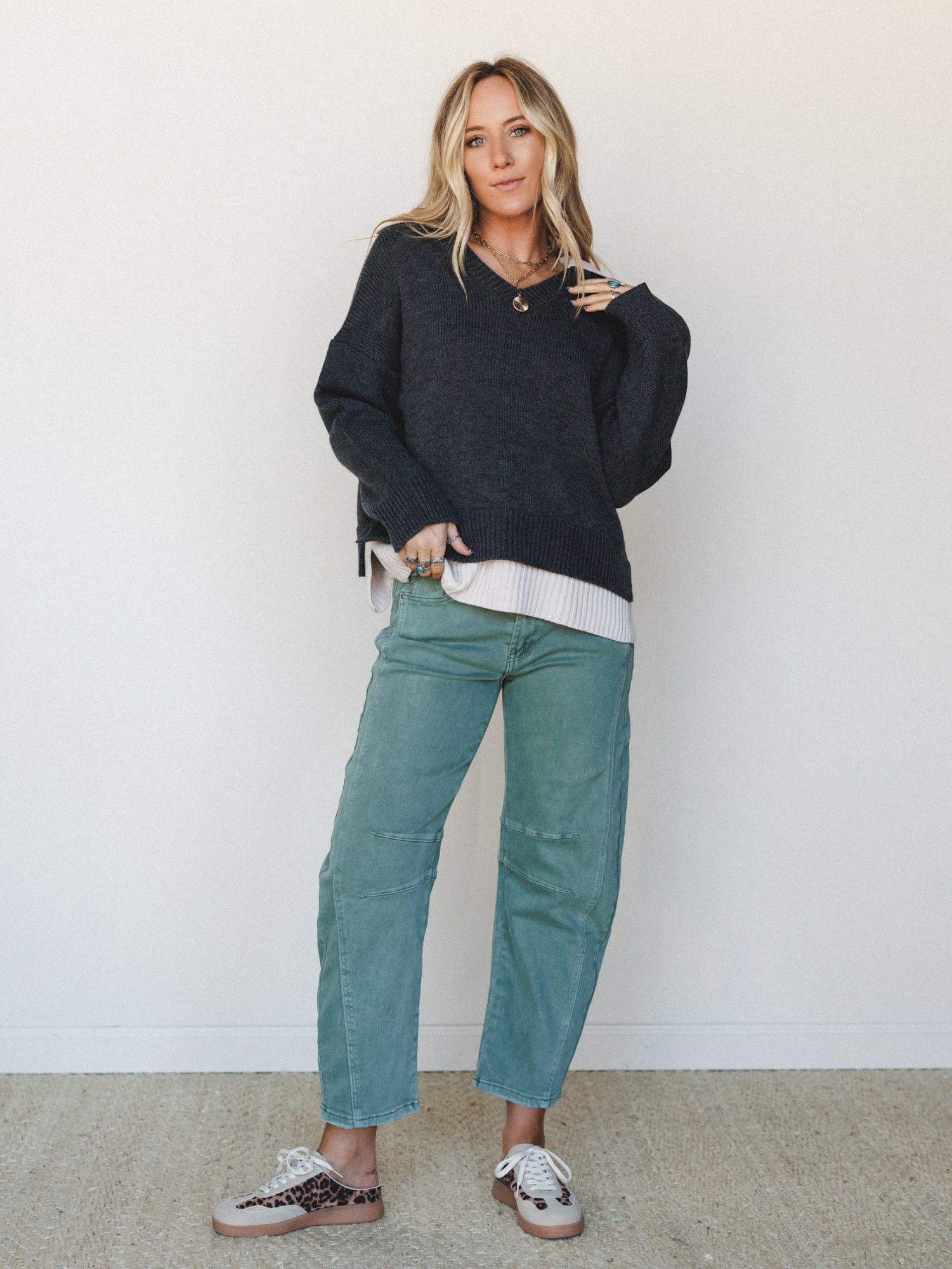 A relaxed, layered fall outfit — oversized charcoal knit sweater over a ribbed cream underlayer, paired with sage green utility-style pants and leopard print sneakers, shot against a clean white wall with an effortlessly put-together, everyday feel.