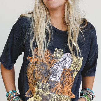 A smiling woman with long blonde hair wears the On The Prowl Distressed Graphic Tee in charcoal, a black mineral-washed shirt with a colorful vintage tiger design. She pairs it with beaded bracelets and stands against a plain light background.