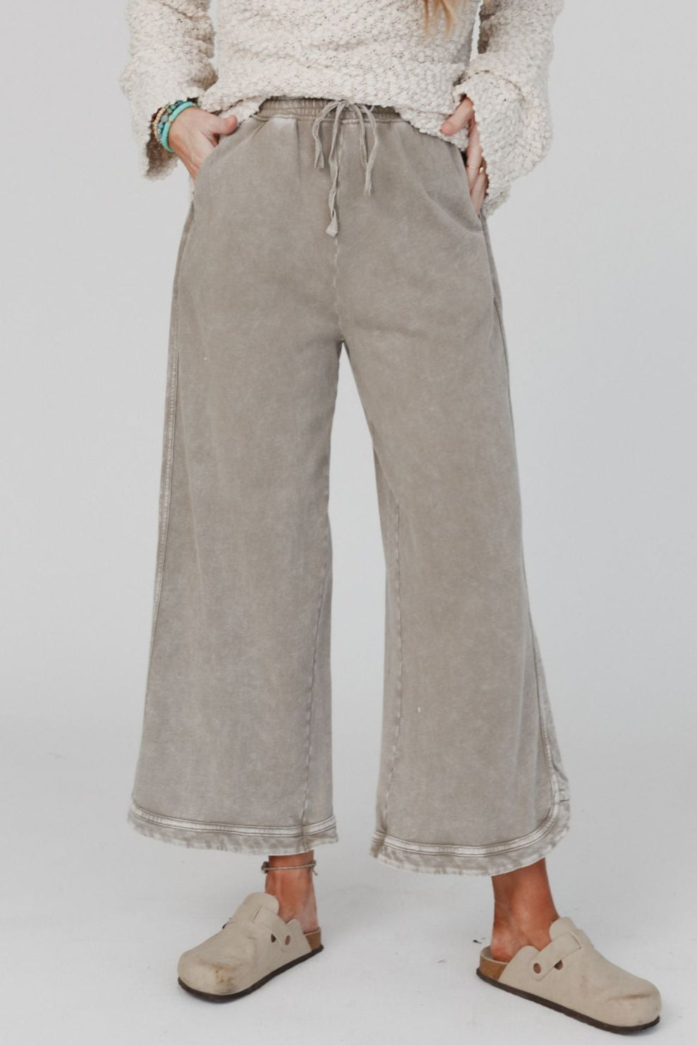 A person stands facing forward, wearing the So Comfy Wide Leg Pant Cropped Length in Mocha with hands in pockets. Only their body from shoulders down is visible. They pair the pants with a textured cream sweater and beige slip-on shoes.