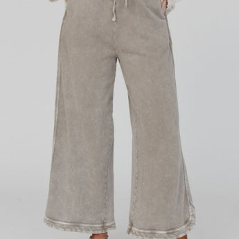 A person stands facing forward, wearing the So Comfy Wide Leg Pant Cropped Length in Mocha with hands in pockets. Only their body from shoulders down is visible. They pair the pants with a textured cream sweater and beige slip-on shoes.