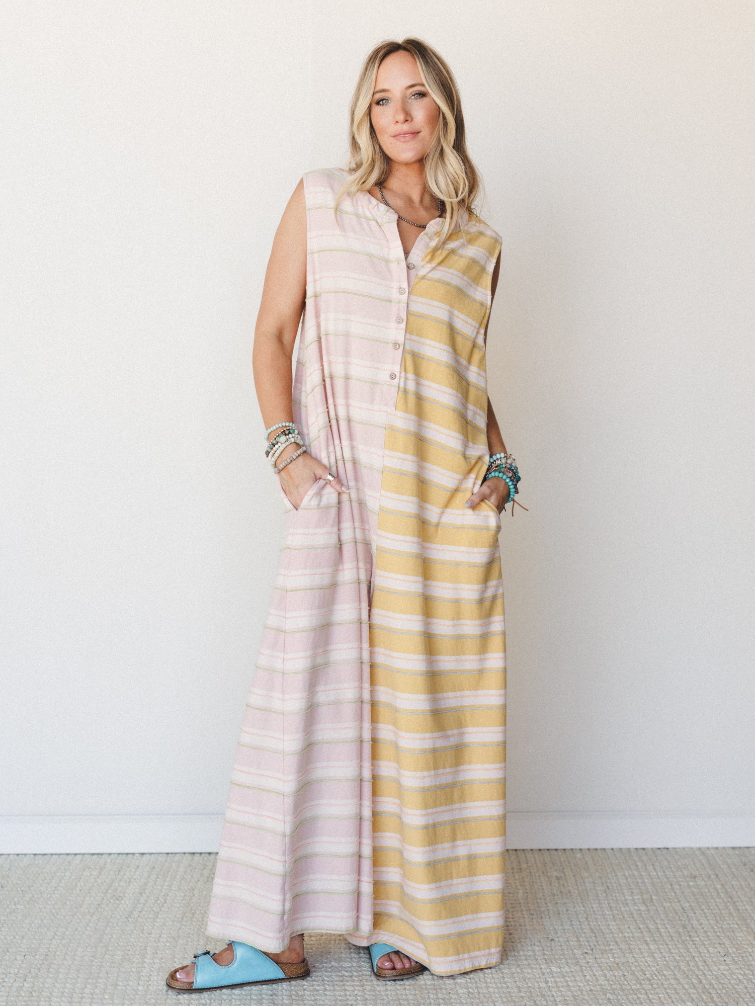 A breezy color-blocked maxi jumpsuit in soft pink and mustard stripes, styled with stacked beaded bracelets and sky blue sandals against a clean studio wall — the kind of outfit that feels like a slow morning somewhere warm.