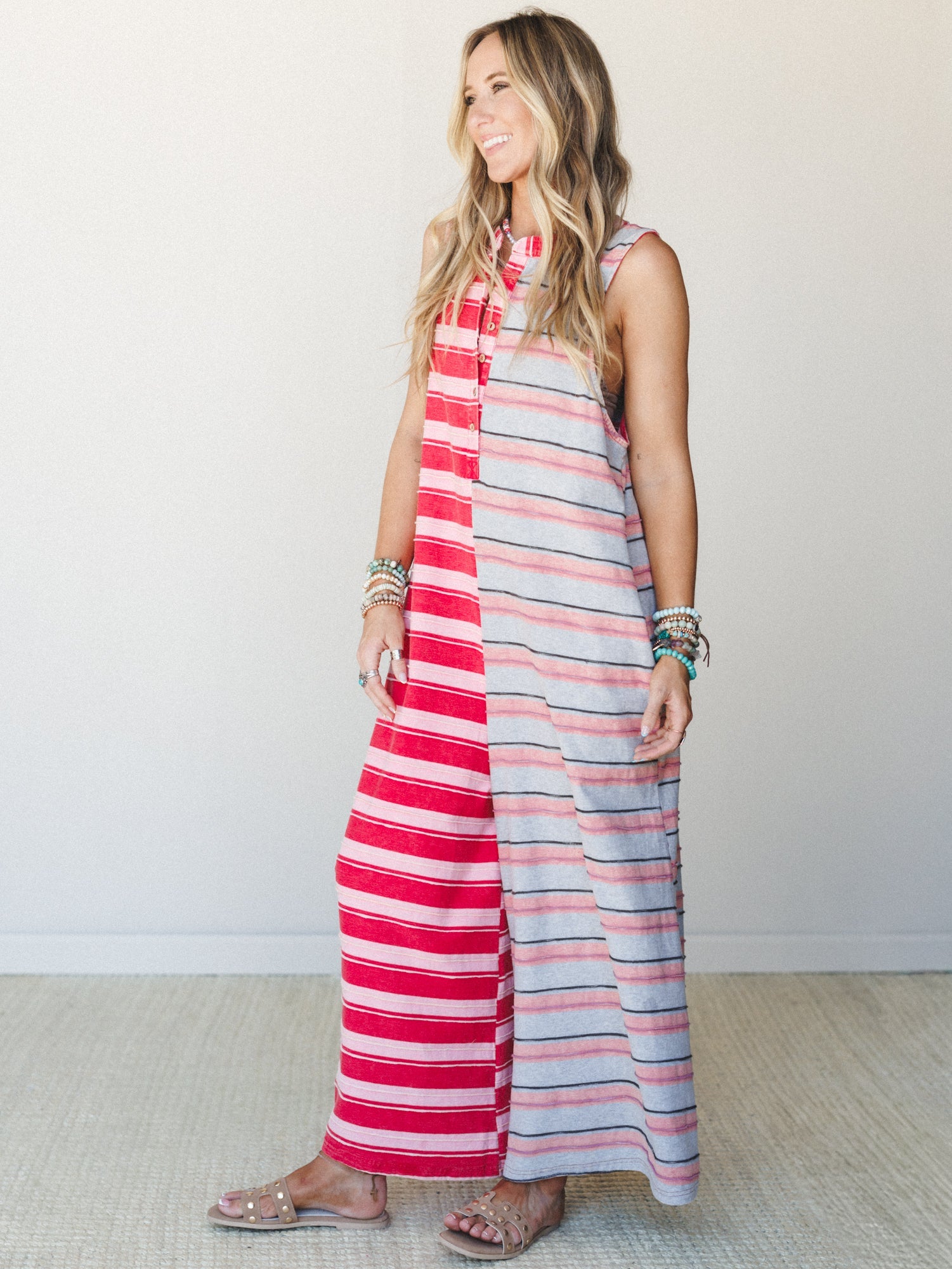 A striped maxi jumpsuit in a bold red-and-grey colorblock split — sleeveless, relaxed, and effortlessly summer — styled with stacked turquoise bracelets and flat sandals against a clean studio wall.