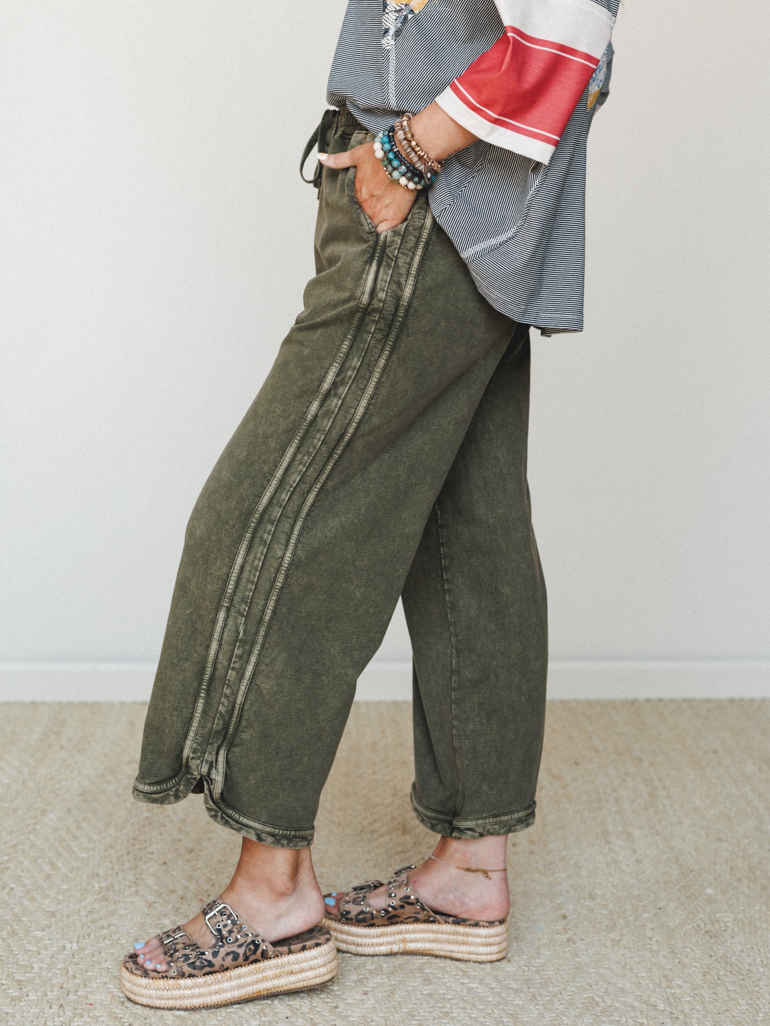 A side-profile shot showing olive mineral-wash wide-leg crop pants styled with a striped colorblock top, stacked bead bracelets, and leopard print platform sandals — relaxed and expressive in the best way.