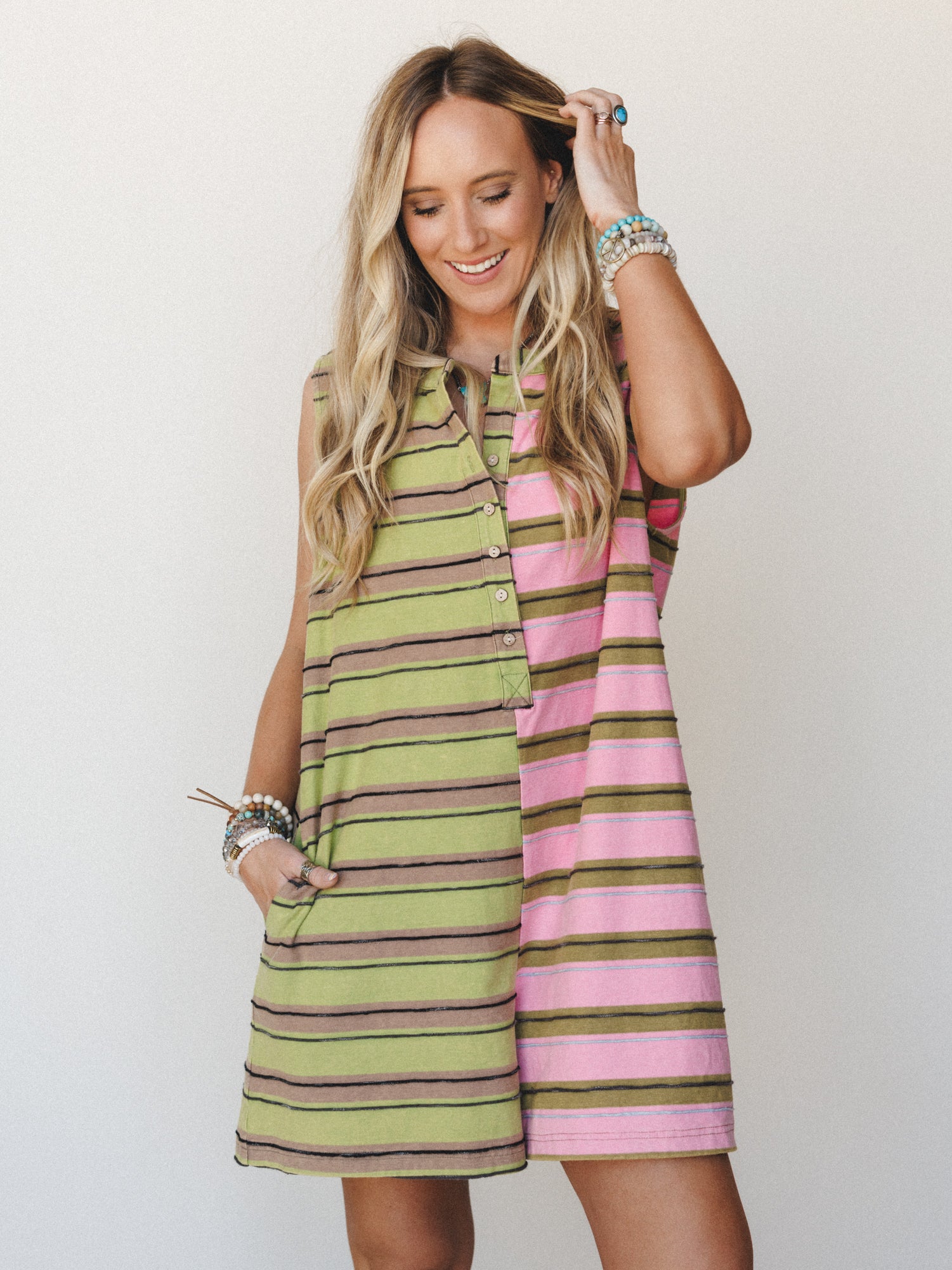 A color-blocked striped mini dress with a relaxed A-line silhouette — split down the center with lime green and pink stripe panels — styled with stacked boho bracelets and a laid-back smile that feels like the first day of a road trip.