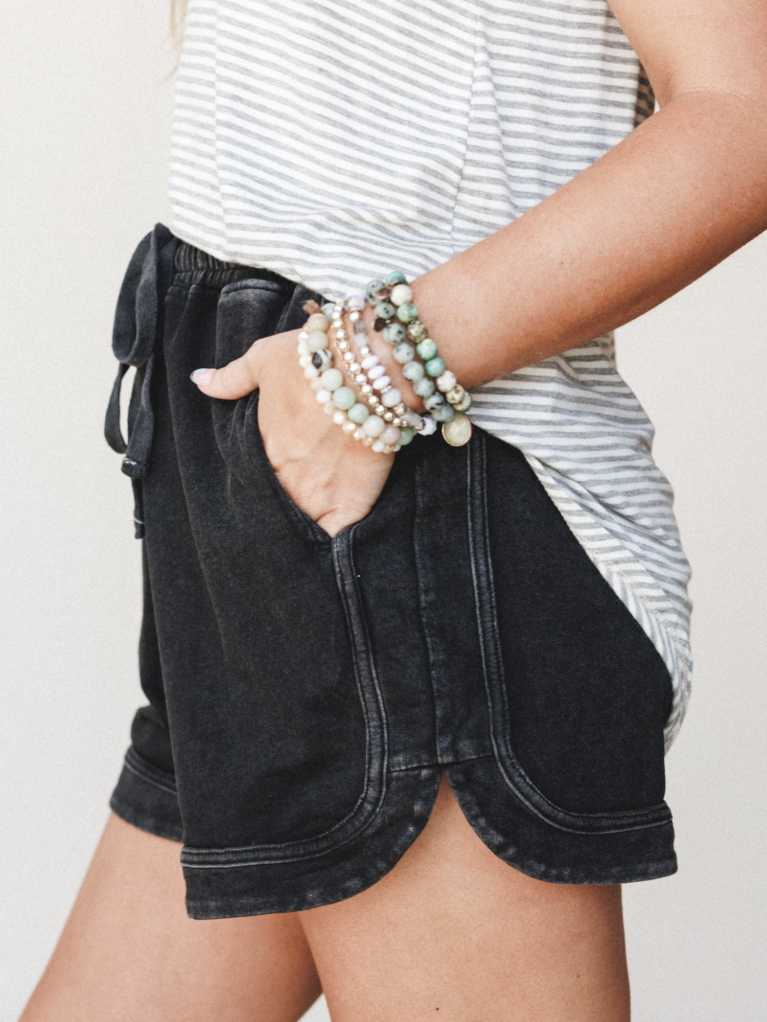 A close-up lifestyle shot of acid-washed black lounge shorts paired with a striped tee, hand tucked in pocket and stacked beaded bracelets adding an easy boho touch.