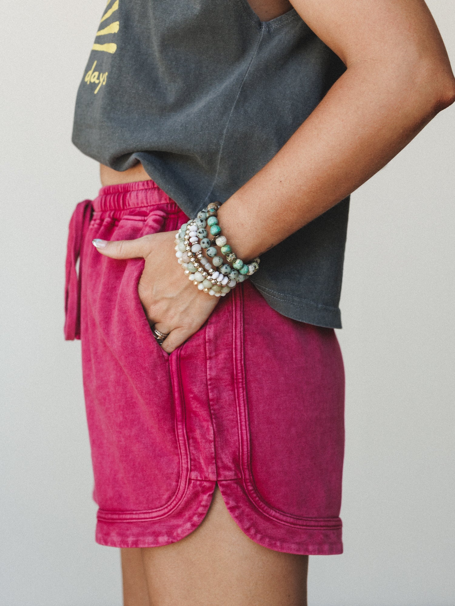A close mid-shot of acid-washed magenta lounge shorts paired with a cropped graphic tee, styled with a stack of beaded stone bracelets — casual and expressive with just enough personality.