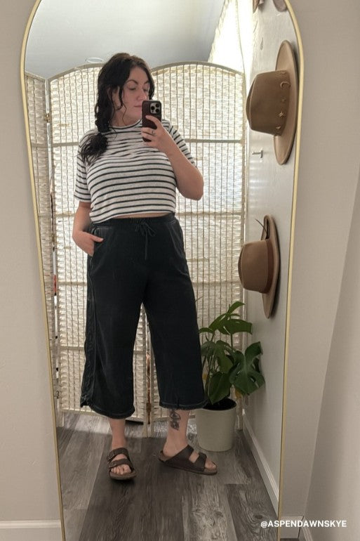A person stands in front of a mirror taking a selfie, wearing a striped t-shirt and the So Comfy Wide Leg Pant Cropped Length - Charcoal. Hats hang on the wall, with a plant nearby and a folding screen in the background.