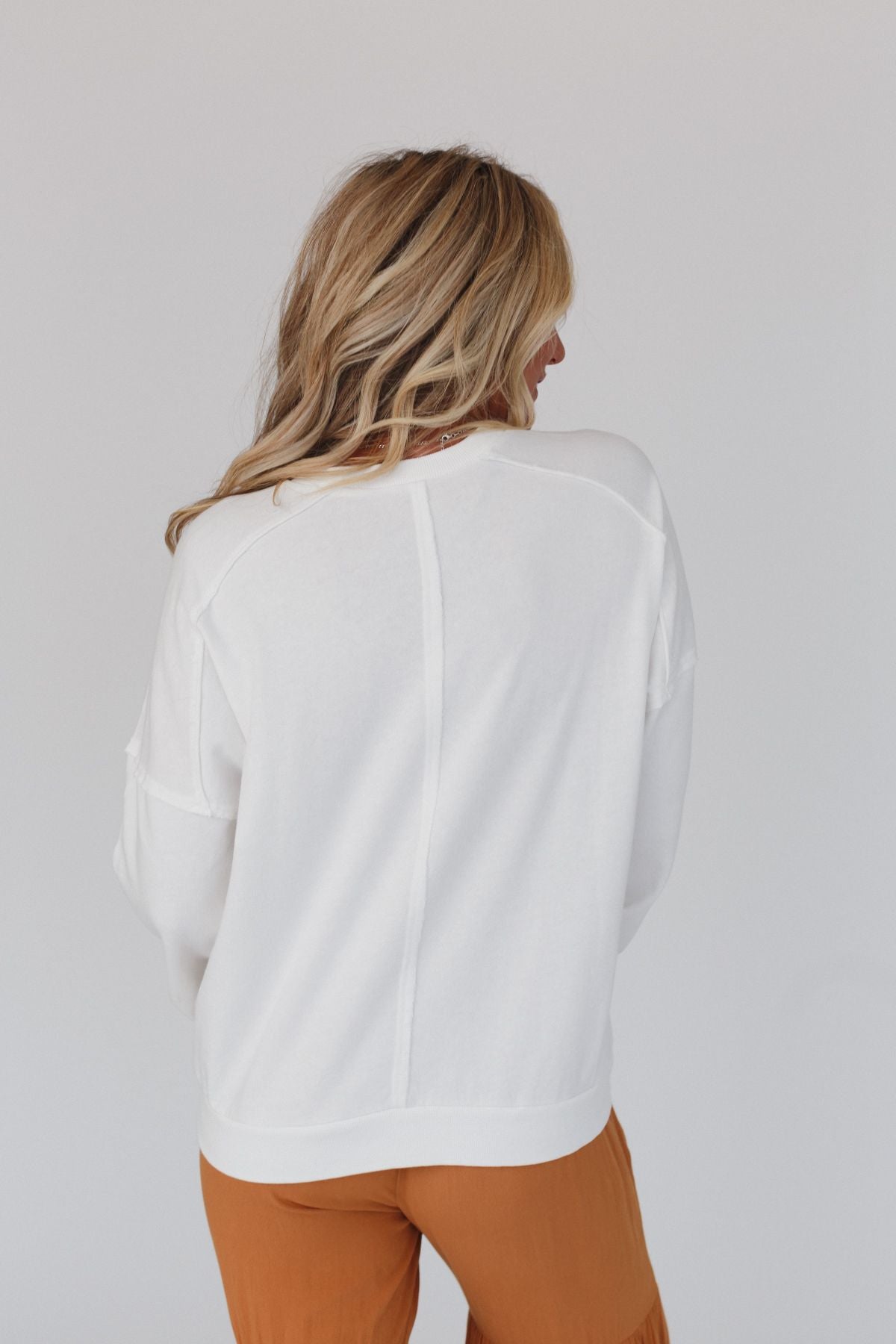 A woman with long, wavy blonde hair faces away from the camera, wearing the Jenny Floral Peace Sign Pullover Top in Off White and rust-colored pants against a plain light background.