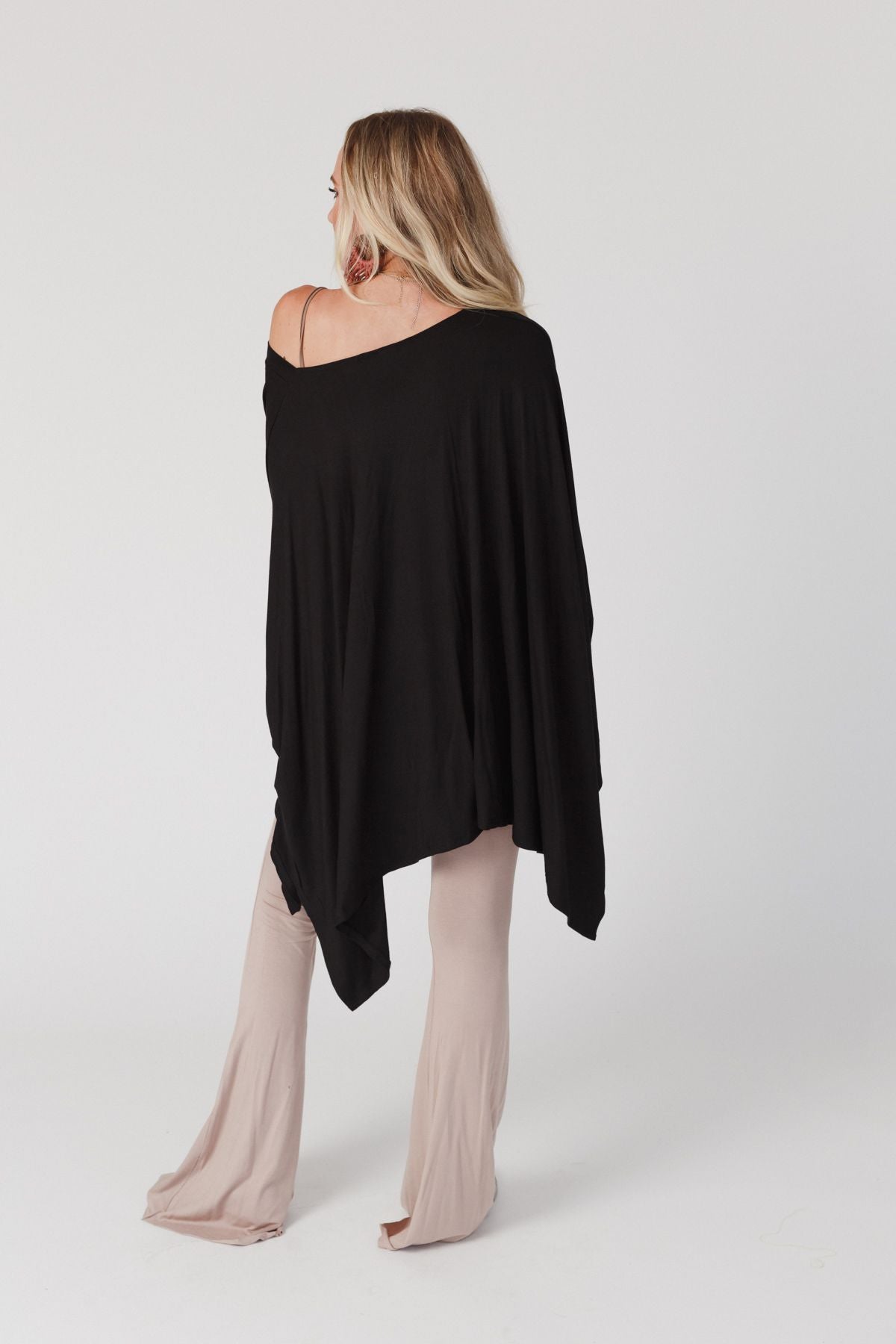 A woman with long blonde hair, seen from behind against a white background, wears The Nest The Wren Top in black—a loose boho top with an asymmetrical, flowy v-neckline over one shoulder—paired with light beige wide-leg pants.